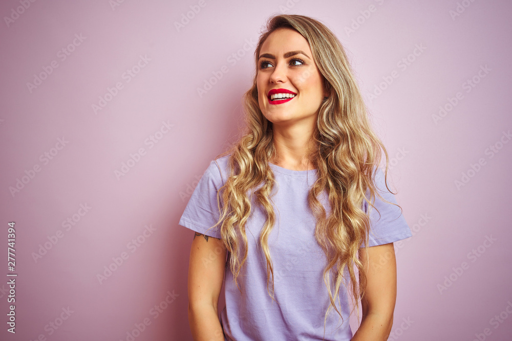 Young beautiful woman wearing purple t-shirt standing over pink isolated background looking away to side with smile on face, natural expression. Laughing confident.