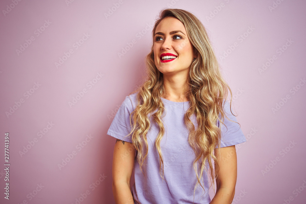 Young beautiful woman wearing purple t-shirt standing over pink isolated background looking away to side with smile on face, natural expression. Laughing confident. Young beautiful woman wearing purple t-shirt standing over pink isolated background looking away to side with smile on face, natural expression. Laughing confident.