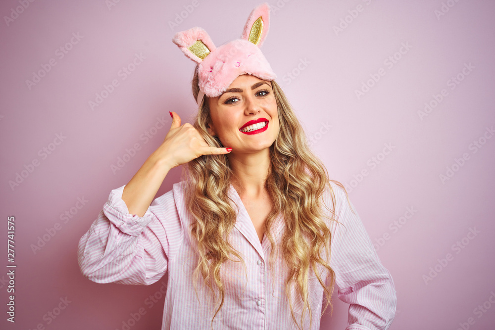 Young beautiful woman wearing pajama and sleep mask over pink isolated background smiling doing phone gesture with hand and fingers like talking on the telephone. Communicating concepts.