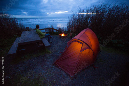 A woman enjoys a beer while sitting on a picnic table next to a camp fire while car camping at a beach side camp site in Kalaloch Campground in the Olympic National Park in Washington State.