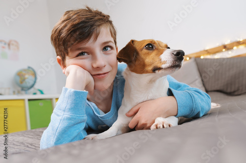 Portrait of a happy boy and his dog breed Jack Russell Terrier, who sit and cuddle on a gray bedspread over the bed and affectionately looking at his owner against the bokeh background of bright light