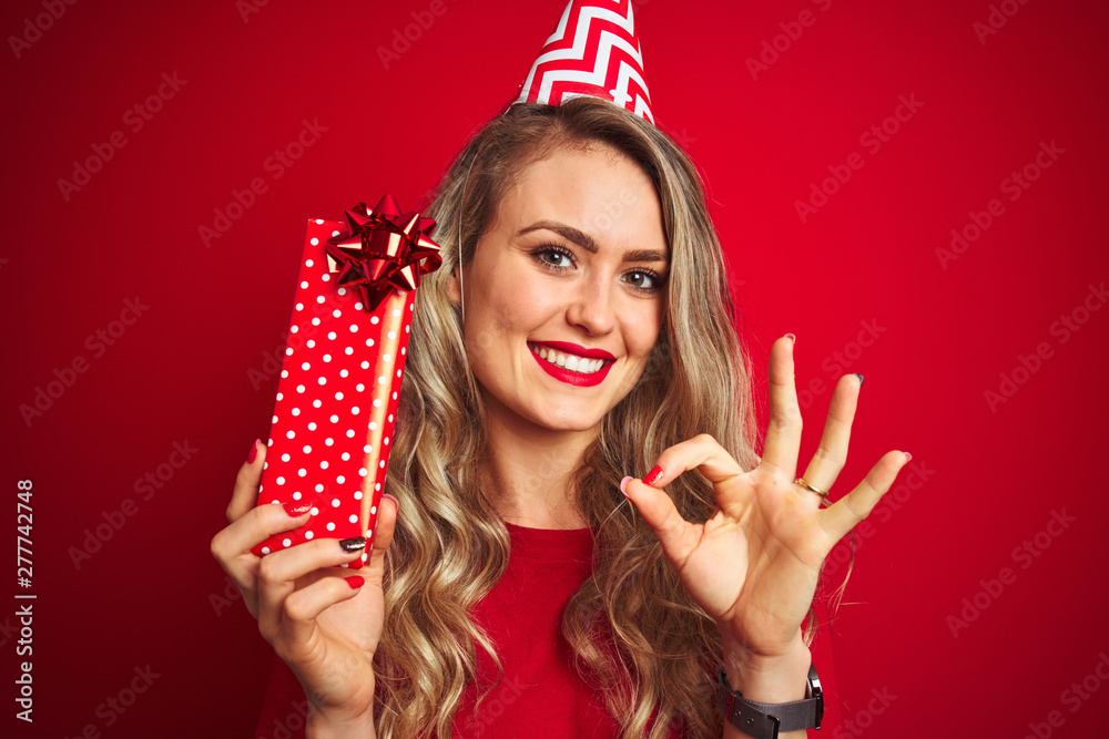 Young beautiful woman holding bitrhday gift over red isolated background doing ok sign with fingers, excellent symbol Young beautiful woman holding bitrhday gift over red isolated background doing ok sign with fingers, excellent symbol