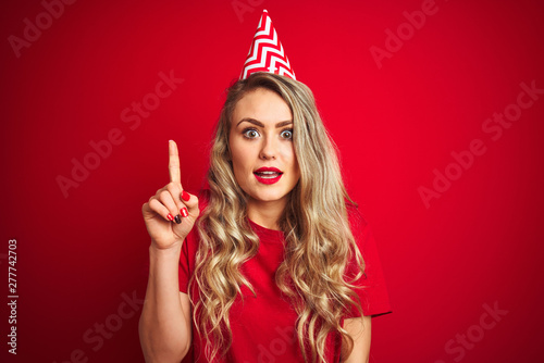Young beautiful woman wearing bitrhday hat over red isolated background surprised with an idea or question pointing finger with happy face, number one