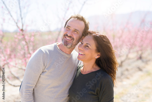 Beautiful middle age senior couple smiling in love at romantic garden of peach trees with pink petals on a sunny day of spring