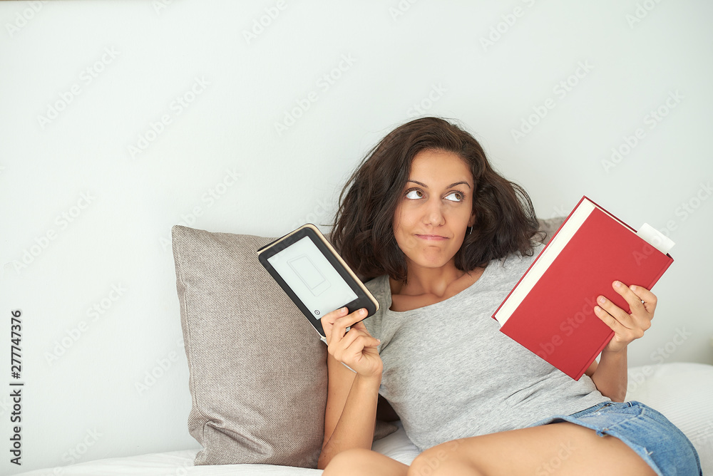 Mujer morena de raza latina sentada en una cama cogiendo un libro en una mano y una tablet en la otra mano, ella tiene una expresion duditativa. El fondo es blanco y liso. Mujer morena de raza latina sentada en una cama cogiendo un libro en una mano y una tablet en la otra mano, ella tiene una expresion duditativa. El fondo es blanco y liso.
