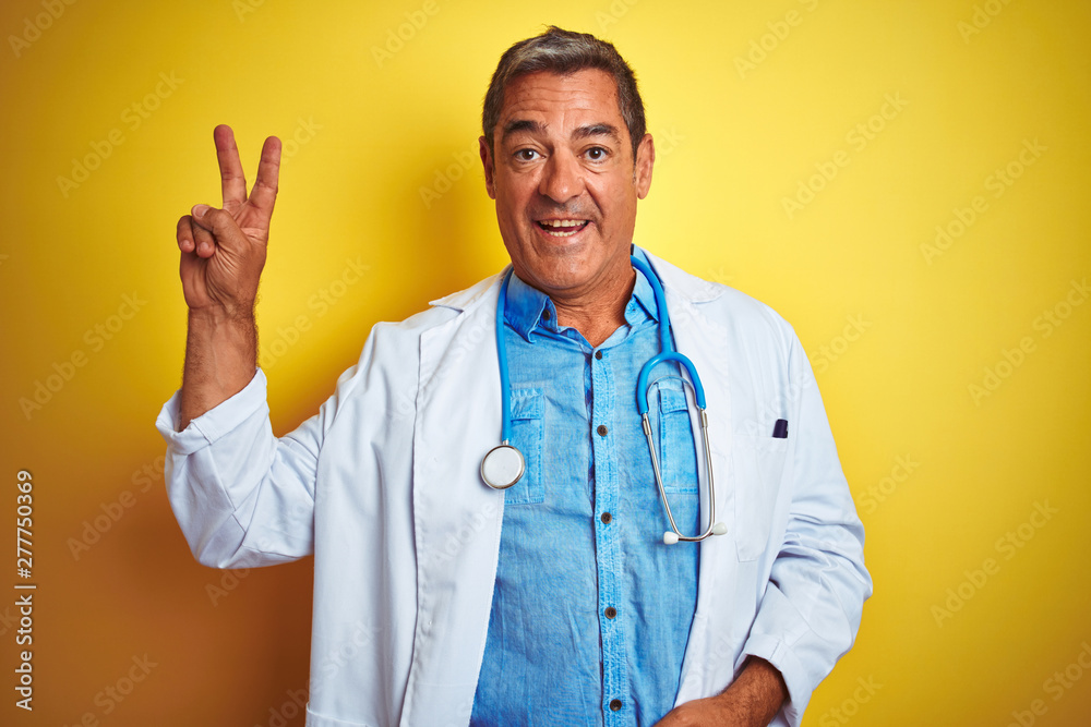 Handsome middle age doctor man wearing stethoscope over isolated yellow background smiling with happy face winking at the camera doing victory sign. Number two. Handsome middle age doctor man wearing stethoscope over isolated yellow background smiling with happy face winking at the camera doing victory sign. Number two.