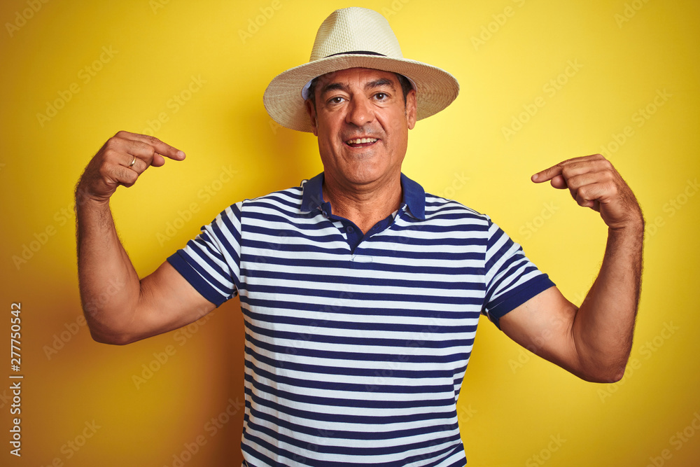 Handsome middle age man wearing striped polo and hat over isolated yellow background looking confident with smile on face, pointing oneself with fingers proud and happy.