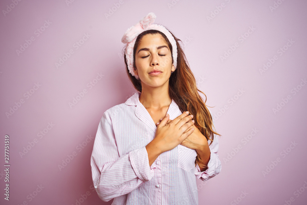 Young beautiful woman wearing pajama standing over pink isolated background smiling with hands on chest with closed eyes and grateful gesture on face. Health concept. Young beautiful woman wearing pajama standing over pink isolated background smiling with hands on chest with closed eyes and grateful gesture on face. Health concept.