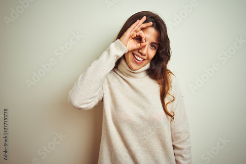 Young beautiful woman wearing winter sweater standing over white isolated background doing ok gesture with hand smiling, eye looking through fingers with happy face. © Krakenimages.com