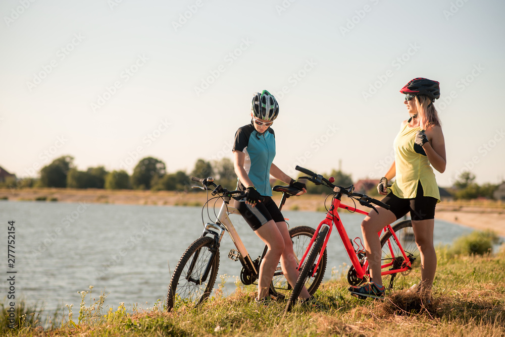 Fototapeta premium Two women cyclists with bikes standing and talking at the lake shore
