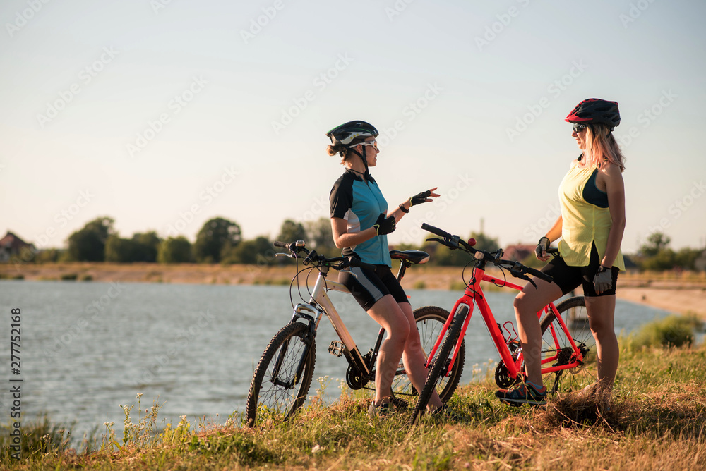 Two women cyclists with bikes standing and talking at the lake shore Two women cyclists with bikes standing and talking at the lake shore