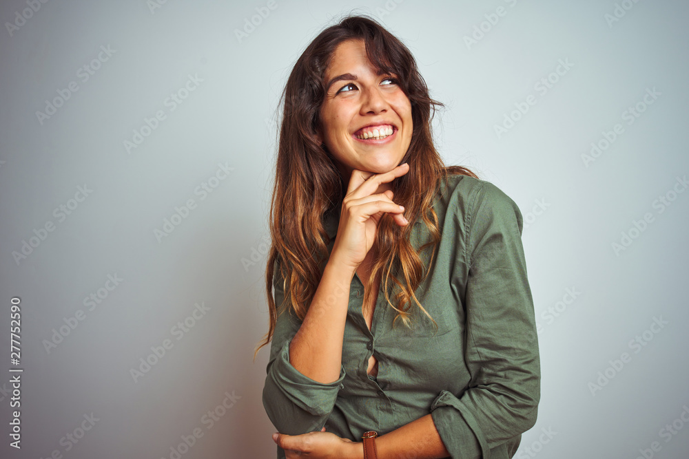 Young beautiful woman wearing green shirt standing over grey isolated background with hand on chin thinking about question, pensive expression. Smiling and thoughtful face. Doubt concept. Young beautiful woman wearing green shirt standing over grey isolated background with hand on chin thinking about question, pensive expression. Smiling and thoughtful face. Doubt concept.