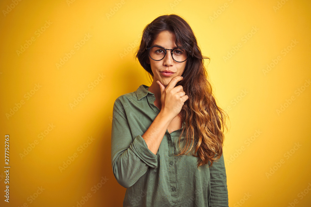 Young beautiful woman wearing green shirt and glasses over yelllow isolated background looking confident at the camera with smile with crossed arms and hand raised on chin. Thinking positive.