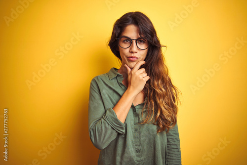 Young beautiful woman wearing green shirt and glasses over yelllow isolated background looking confident at the camera with smile with crossed arms and hand raised on chin. Thinking positive.