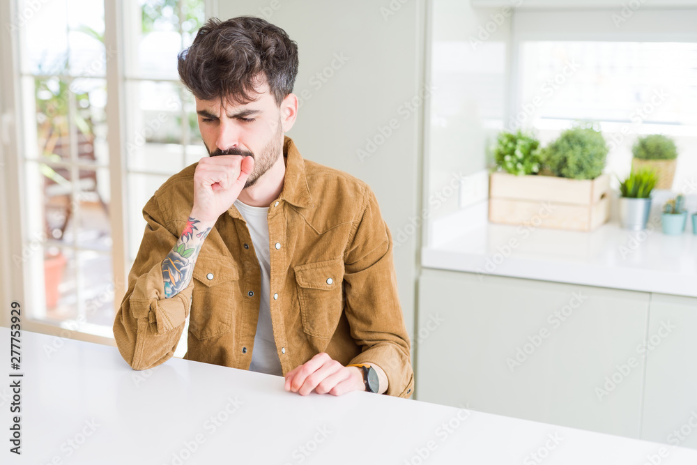 Young man wearing casual jacket sitting on white table feeling unwell and coughing as symptom for cold or bronchitis. Healthcare concept.
