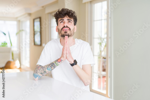 Young man wearing casual t-shirt sitting on white table begging and praying with hands together with hope expression on face very emotional and worried. Asking for forgiveness. Religion concept.