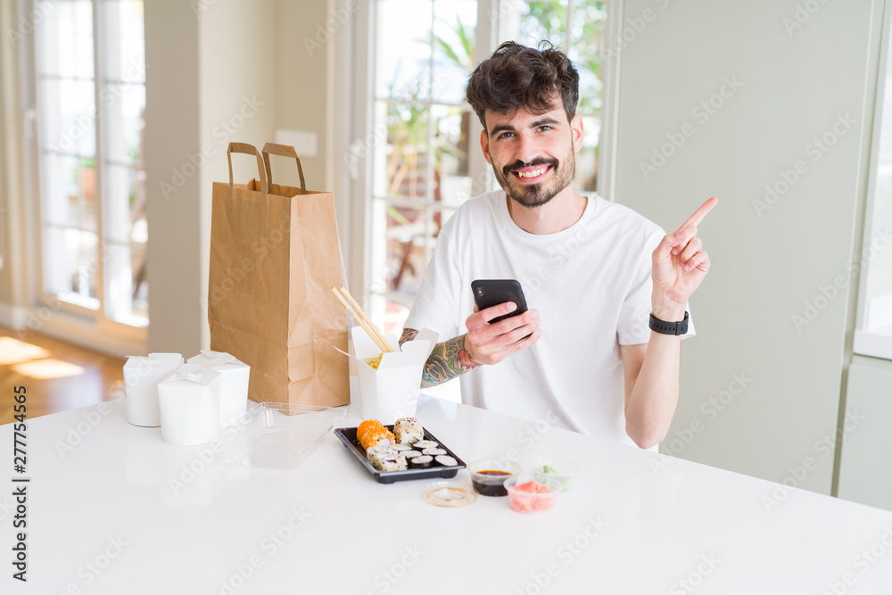 Young man eating asian sushi from home delivery and ordering food using smartphone app very happy pointing with hand and finger to the side Young man eating asian sushi from home delivery and ordering food using smartphone app very happy pointing with hand and finger to the side