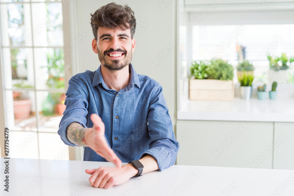 Young man wearing casual shirt sitting on white table smiling friendly offering handshake as greeting and welcoming. Successful business.