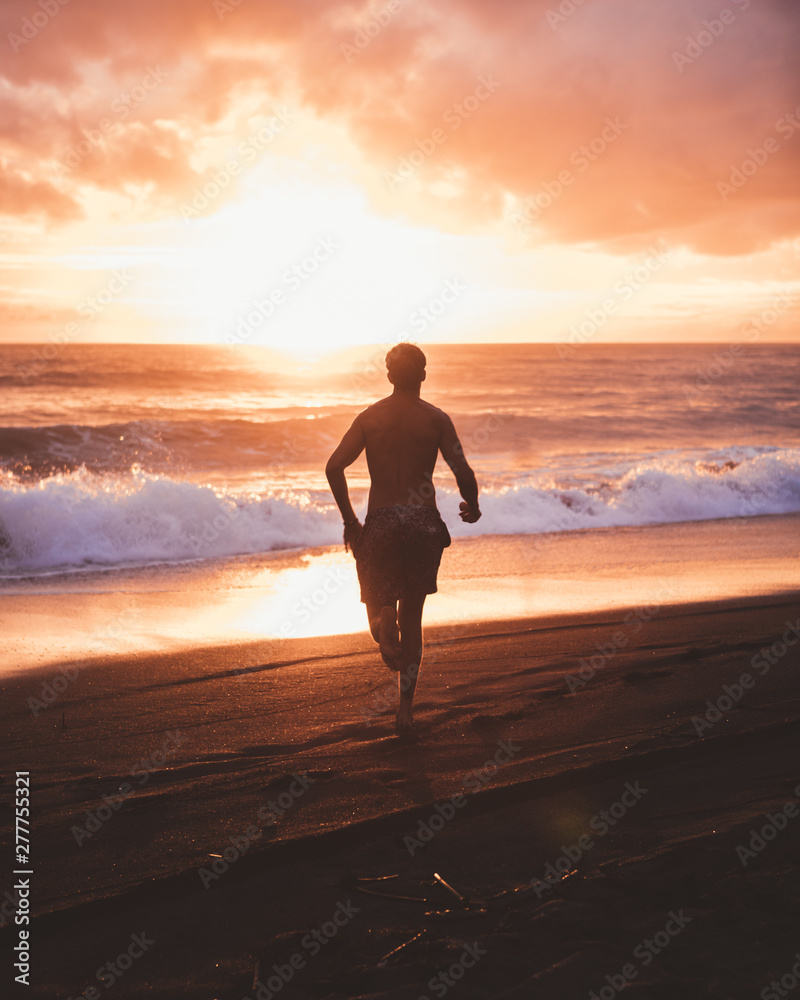Back view of man running on coastline against powerful ocean waves and majestic sunset sky, Bali Back view of man running on coastline against powerful ocean waves and majestic sunset sky, Bali