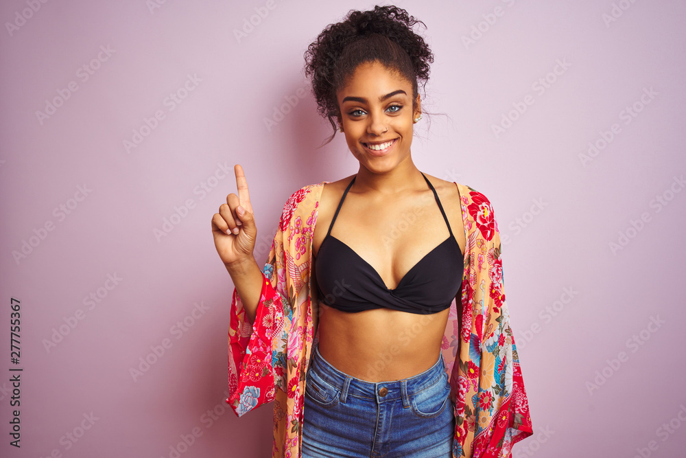 American woman on holiday wearing bikini and colorful dress over isolated pink background showing and pointing up with finger number one while smiling confident and happy. American woman on holiday wearing bikini and colorful dress over isolated pink background showing and pointing up with finger number one while smiling confident and happy.