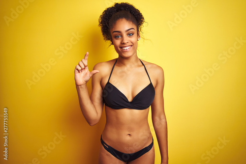 African american woman on vacation wearing bikini standing over isolated yellow background smiling and confident gesturing with hand doing small size sign with fingers looking and the camera. 