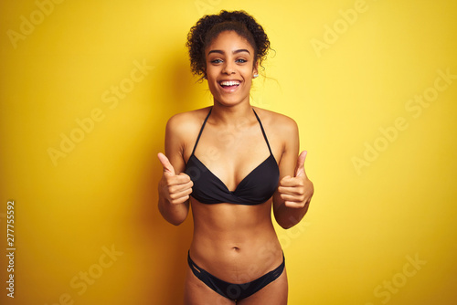 African american woman on vacation wearing bikini standing over isolated yellow background success sign doing positive gesture with hand  thumbs up smiling and happy. Cheerful expression 