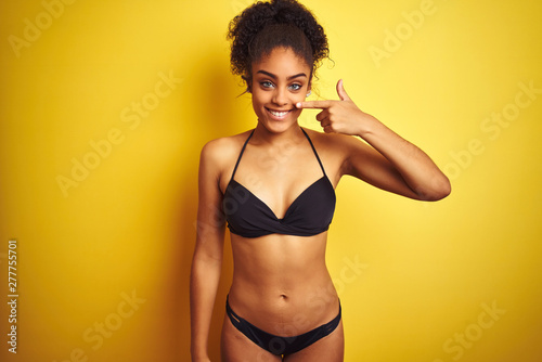 African american woman on vacation wearing bikini standing over isolated yellow background Pointing with hand finger to face and nose, smiling cheerful. Beauty concept