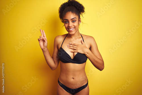 African american woman on vacation wearing bikini standing over isolated yellow background smiling swearing with hand on chest and fingers up, making a loyalty promise oath