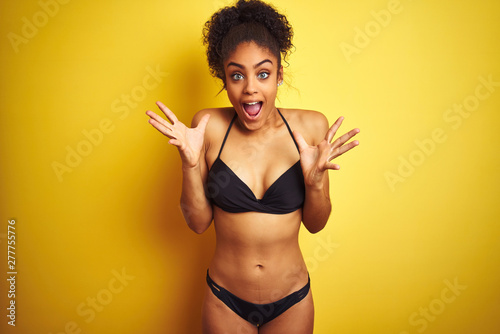 African american woman on vacation wearing bikini standing over isolated yellow background celebrating crazy and amazed for success with arms raised and open eyes screaming excited. Winner concept © Krakenimages.com