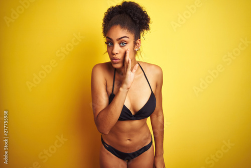 African american woman on vacation wearing bikini standing over isolated yellow background hand on mouth telling secret rumor, whispering malicious talk conversation