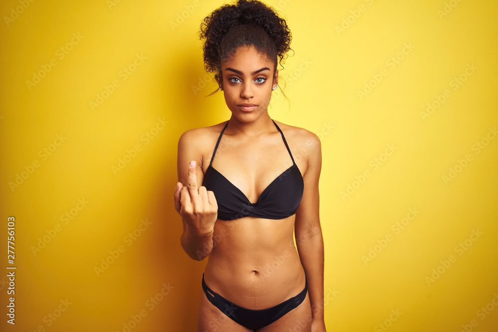 African american woman on vacation wearing bikini standing over isolated yellow background Showing middle finger, impolite and rude fuck off expression African american woman on vacation wearing bikini standing over isolated yellow background Showing middle finger, impolite and rude fuck off expression