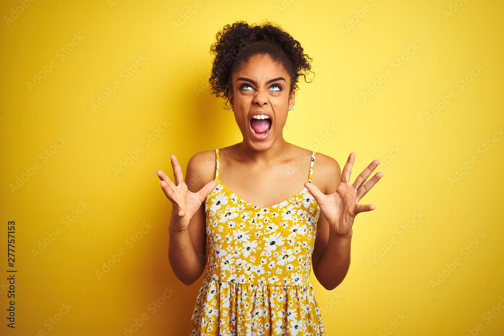 African american woman wearing casual floral dress standing over isolated yellow background crazy and mad shouting and yelling with aggressive expression and arms raised. Frustration concept. African american woman wearing casual floral dress standing over isolated yellow background crazy and mad shouting and yelling with aggressive expression and arms raised. Frustration concept.