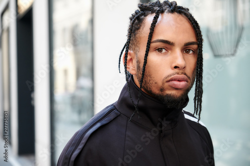 Young ethnic man with braided hair wearing black sports suit looking at camera on urban background photo