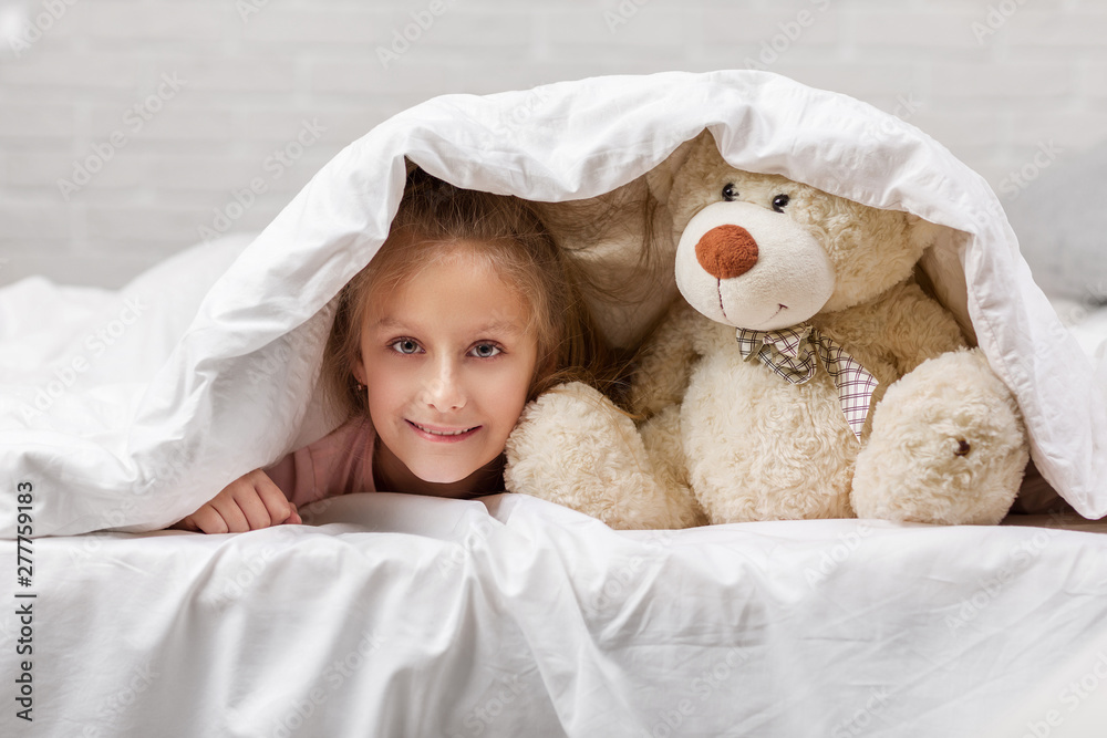 adorable little child girl playing with teddy bear in bed in morning. baby with a teddy bear under the covers