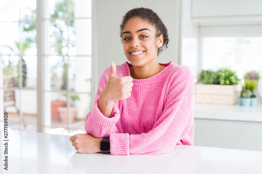 Beautiful african american woman with afro hair wearing casual pink sweater doing happy thumbs up gesture with hand. Approving expression looking at the camera showing success. Beautiful african american woman with afro hair wearing casual pink sweater doing happy thumbs up gesture with hand. Approving expression looking at the camera showing success.