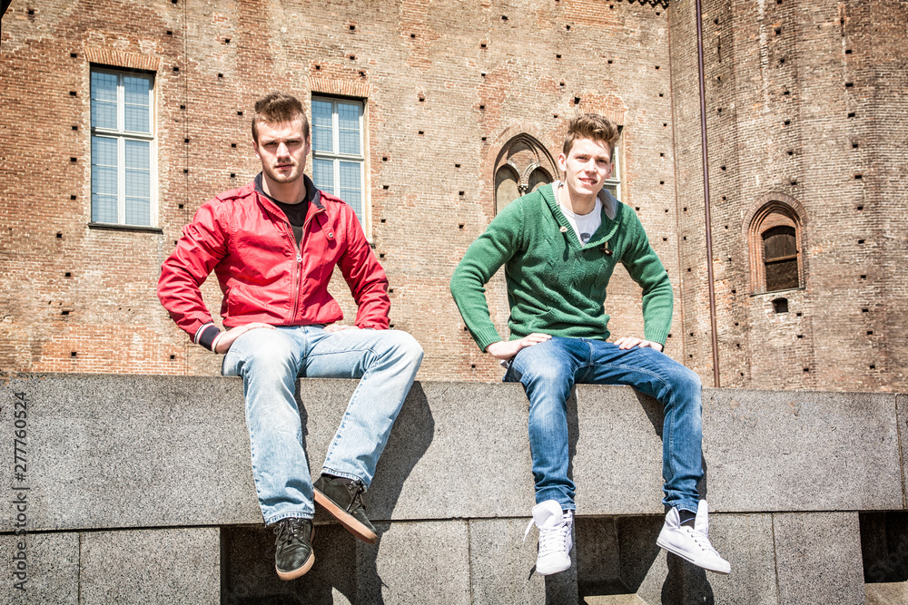 Two young men sitting on low wall in urban setting in European city. Summertime