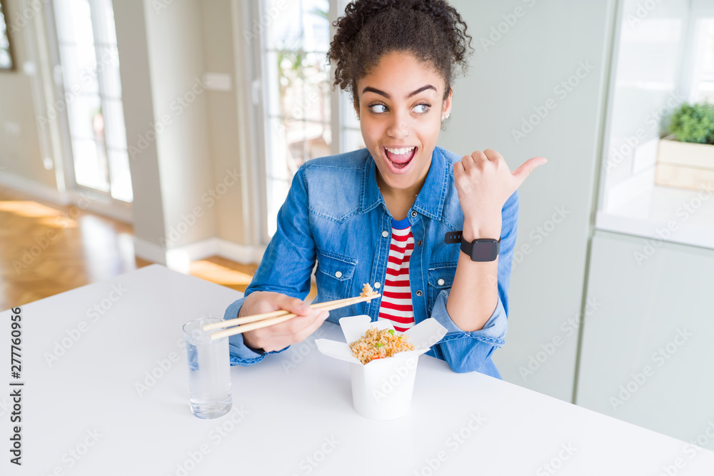 Young african american woman eating asian noodles from delivery box pointing and showing with thumb up to the side with happy face smiling Young african american woman eating asian noodles from delivery box pointing and showing with thumb up to the side with happy face smiling