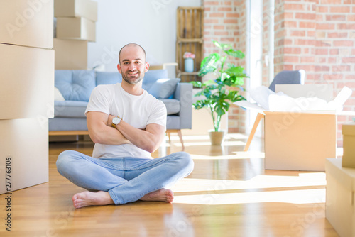 Young bald man sitting on the floor around cardboard boxes moving to a new home happy face smiling with crossed arms looking at the camera. Positive person.