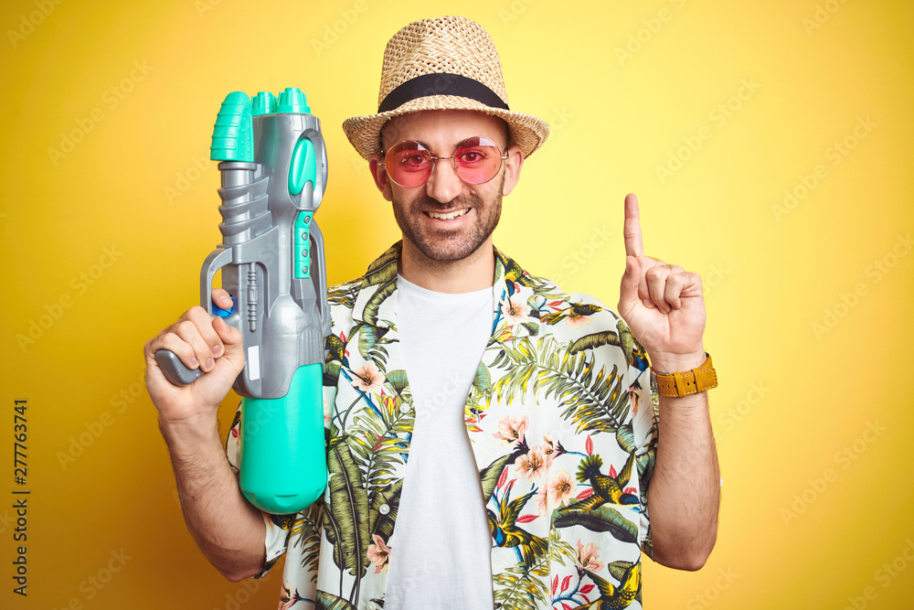Young man wearing hawaiian flowers shirt holding water gun over yellow isolated background surprised with an idea or question pointing finger with happy face, number one