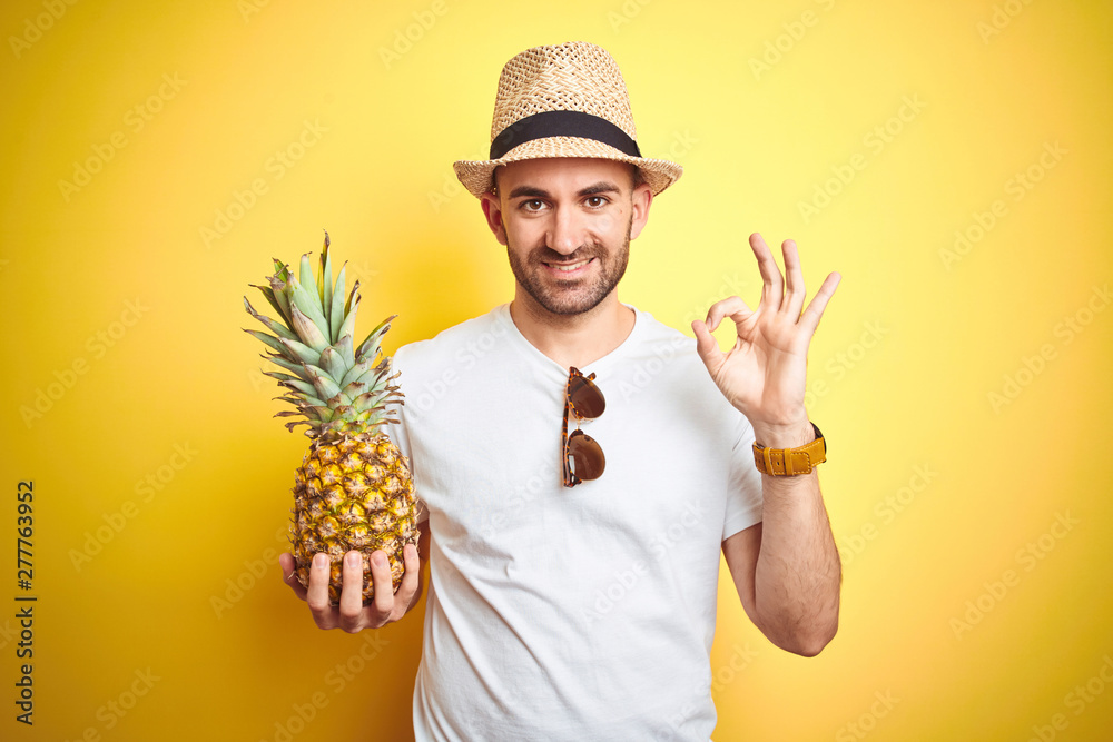 Young man wearing summer hat and holding pineapple over yellow background doing ok sign with fingers, excellent symbol Young man wearing summer hat and holding pineapple over yellow background doing ok sign with fingers, excellent symbol
