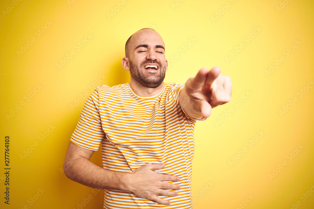 Young bald man with beard wearing casual striped t-shirt over yellow isolated background laughing at you, pointing finger to the camera with hand over body, shame expression Young bald man with beard wearing casual striped t-shirt over yellow isolated background laughing at you, pointing finger to the camera with hand over body, shame expression