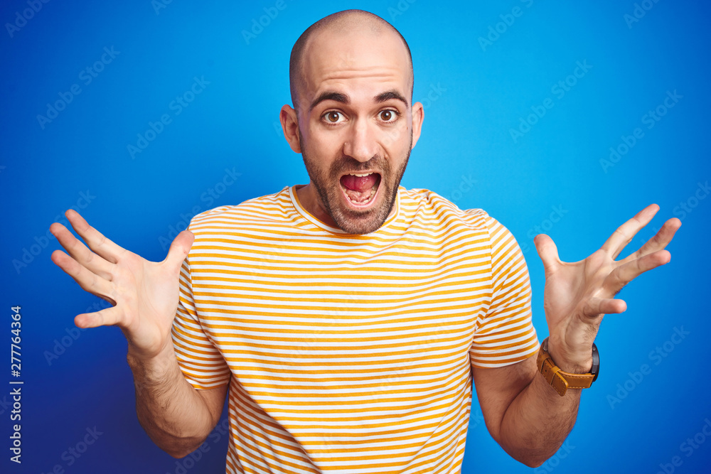 Young bald man with beard wearing casual t-shirt over blue isolated background celebrating crazy and amazed for success with arms raised and open eyes screaming excited. Winner concept