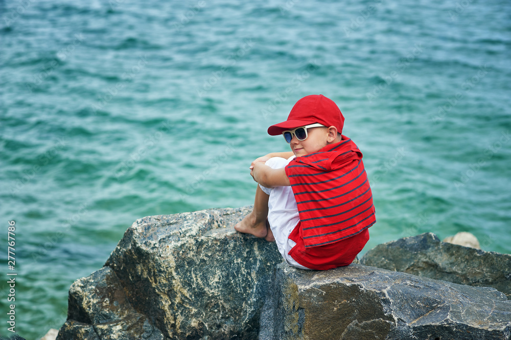 Fashionable handsome boy on the sea coast . Rest and travel . The child sits on a stone in light trousers . Fashionable handsome boy on the sea coast . Rest and travel . The child sits on a stone in light trousers .