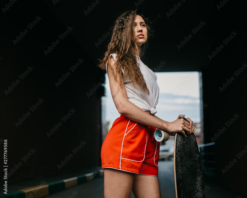Beautiful skater woman wearing red shorts riding on her longboard in the sunset city Beautiful skater woman wearing red shorts riding on her longboard in the sunset city