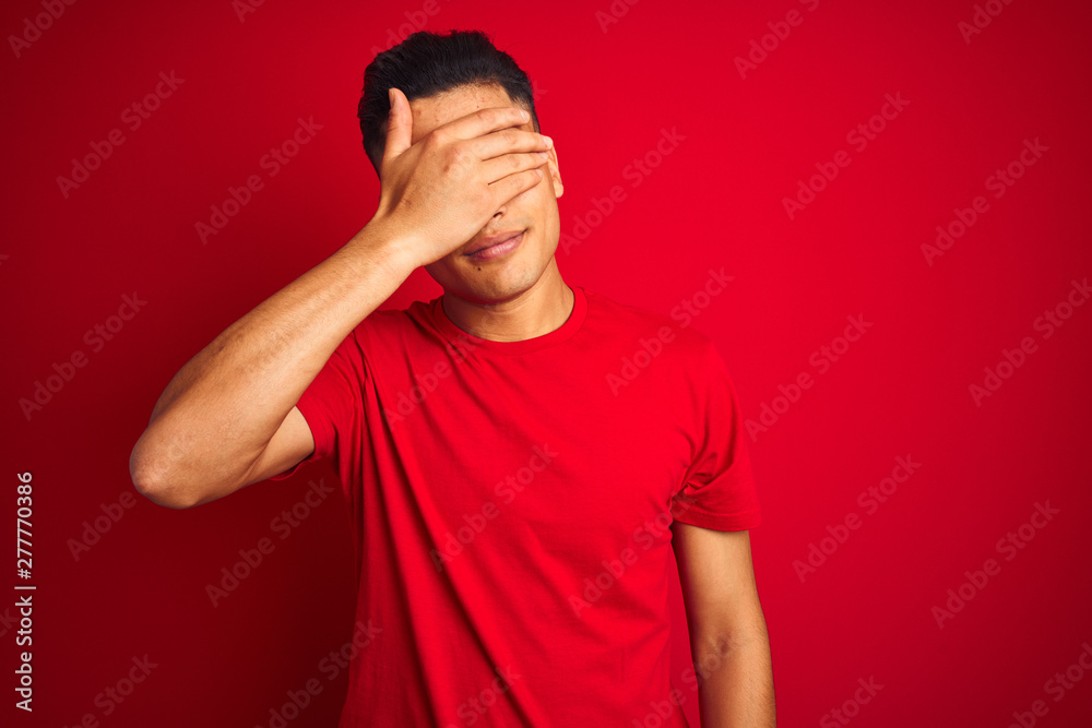 Young brazilian man wearing t-shirt standing over isolated red background covering eyes with hand, looking serious and sad. Sightless, hiding and rejection concept Young brazilian man wearing t-shirt standing over isolated red background covering eyes with hand, looking serious and sad. Sightless, hiding and rejection concept