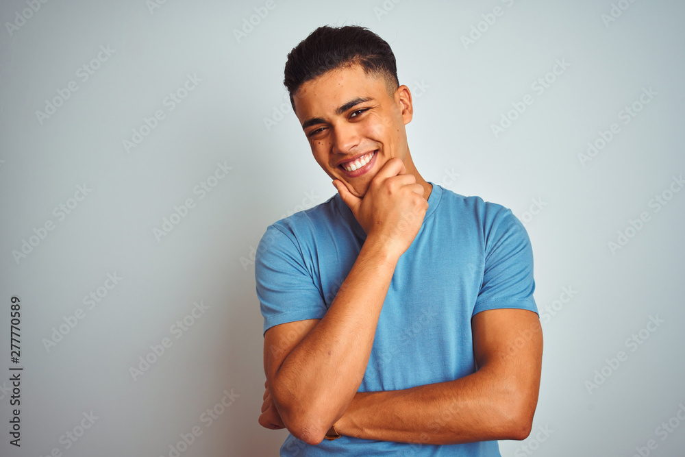 Young brazilian man wearing blue t-shirt standing over isolated white background looking confident at the camera smiling with crossed arms and hand raised on chin. Thinking positive. Young brazilian man wearing blue t-shirt standing over isolated white background looking confident at the camera smiling with crossed arms and hand raised on chin. Thinking positive.