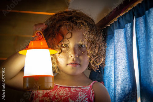 Cute little child with curly hair holding glowing flashlight and looking at camera at home photo