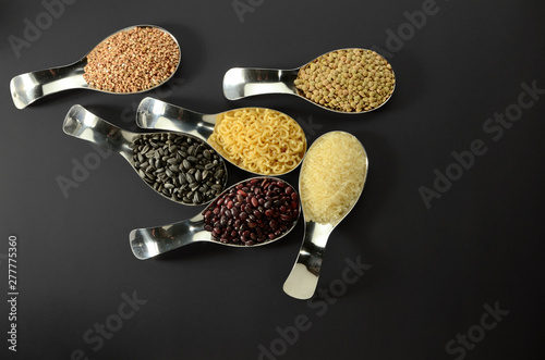 Rice, buckwheat, beans, lentils, seeds, pasta in spoons on a dark background.
