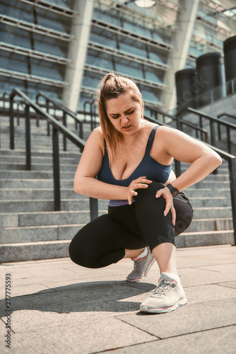 Fototapeta Naklejka Na Ścianę i Meble -  Feeling pain after workout. Young plus size woman touching her knee and feeling pain after sport training while standing on stairs.