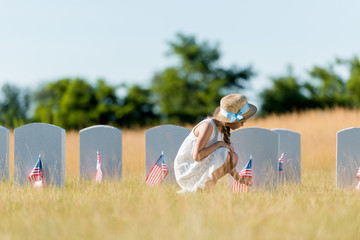 selective focus of kid in dress and straw hat sitting near headstone with american flag in graveyard